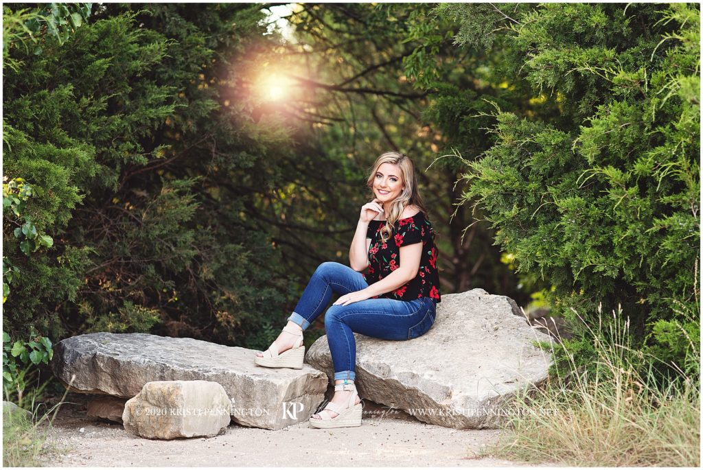 Senior girl in jeans and black and red floral top sitting on rocks at sunset